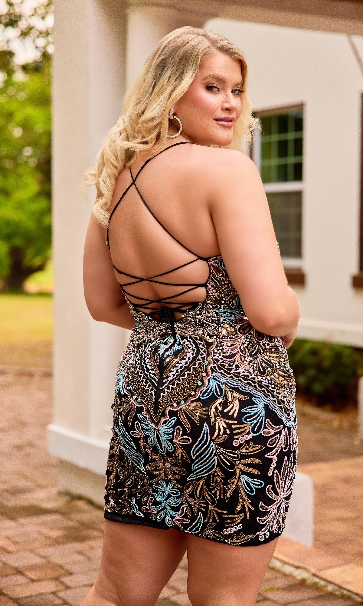 A woman in the Primavera 14090 Plus-Size Short Lace-Up Hoco Dress stands outdoors near a white building, glancing over her shoulder.